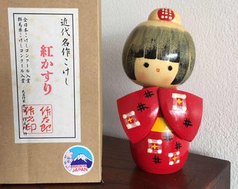 Może przedstawiać: Tradycyjna japońska lalka Kokeshi w czerwono-czarnym kimono. Lalka ma okrągłą głowę z czarnymi włosami i malowaną twarzą. Lalka stoi obok pudełka z japońskim tekstem i naklejką "Made in Japan".
