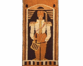 May include: A woven wall hanging with a brown, orange, and white design of a person in a military uniform holding a drum. The design is framed by a brown border with a geometric pattern.