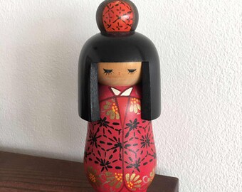 Może przedstawiać: Drewniana japońska lalka Kokeshi z czarną głową i włosami oraz czerwonym kimono ozdobionym czarnymi i złotymi wzorami kwiatowymi. Lalka ma zamknięte oczy i stoi na drewnianej powierzchni.