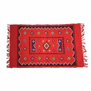 May include: Red wool rug with a geometric pattern in shades of red, green, white, and black. The rug has fringe on all four sides.