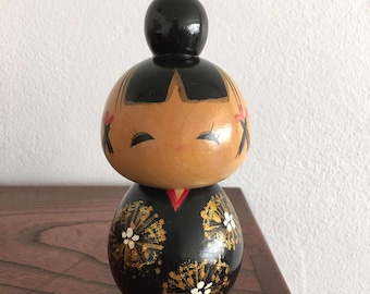 Puede incluir: Una muñeca Kokeshi japonesa de madera con un moño negro y una cara pintada. El cuerpo de la muñeca es negro con diseños florales dorados y blancos. La muñeca tiene un cuello rojo y está sobre una superficie de madera.