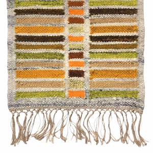 Puede incluir: Una alfombra rectangular tejida con un patrón geométrico de rayas horizontales en tonos de verde, marrón, naranja y amarillo. La alfombra tiene un borde gris y flecos a lo largo del borde inferior. La alfombra está hecha de un material texturizado.