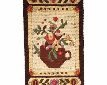 Op de afbeelding: Een rechthoekige, geweven wandtapijt met een bloemenontwerp. Een bruine vaas gevuld met kleurrijke bloemen op een crèmekleurige achtergrond. De rand is bruin met bloemaccenten en een franje aan de onderkant. Ongeveer 60 cm lang.