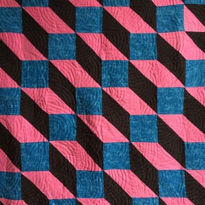 May include: A patterned quilt with a repeating geometric design. The design features squares and diagonal stripes in black, bright pink, and blue. The quilt has a textured appearance with visible stitching.