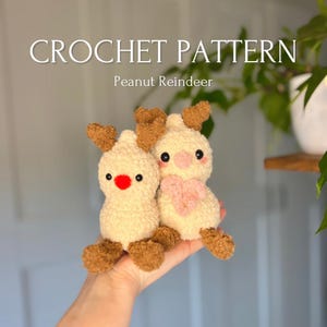Puede incluir: Dos juguetes de reno de cacahuete de crochet. Uno es crema con una nariz roja y cuernos marrones. El otro es crema con una nariz rosa y una bufanda rosa y cuernos marrones. Los juguetes se sostienen en una mano.