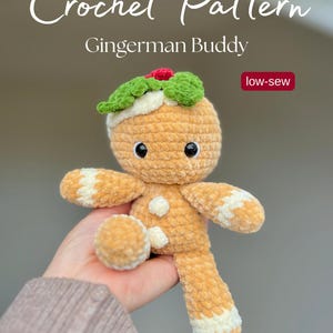 Könnte beinhalten: Ein gehäkelter Lebkuchenmann-Plüsch mit einem grünen und roten Stechpalmenblatt-Design auf dem Kopf. Der Plüsch hat schwarze Augen und weiße Akzente an Armen und Beinen. Der Text "Crochet Pattern" und "Gingerman Buddy" sind sichtbar.