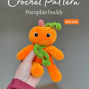 Könnte beinhalten: Eine gehäkelte Kürbis-Buddy-Puppe in Orange mit einem grünen Schal und Stiel. Die Puppe hat schwarze Knopfaugen und wird in einer Hand gehalten. Der Text auf dem Bild lautet "Crochet Pattern Pumpkin buddy" und "low-sew".
