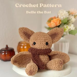 Könnte beinhalten: Ein hellbraunes Häkel-Fledermaus-Plüschtier mit großen Ohren, schwarzen Sicherheitsaugen und einem dunkelbraunen Schal. Der Text Crochet Pattern Belle the Bat ist über dem Plüschtier zu sehen. Das Spielzeug hat eine strukturierte, flauschige Optik.