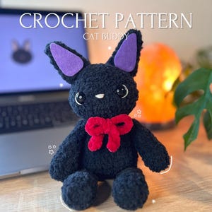 Könnte beinhalten: Häkelanleitung für einen schwarzen Katzenfreund mit lila Ohren und einer roten Schleife. Die Katze sitzt auf einer Holzoberfläche.