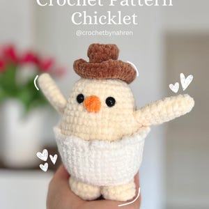 Könnte beinhalten: Eine gehäkelte Chicklet-Puppe mit braunem Hut, orangefarbenem Schnabel und schwarzen Augen. Die Puppe trägt eine weiße Windel und hat ausgestreckte Arme. Der Text "Crochet Pattern Chicklet" und der Handle des Verkäufers sind sichtbar.
