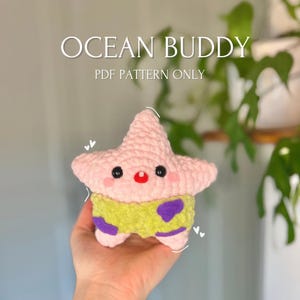 Op de afbeelding: Een roze gehaakte zeester met een groen en paars gehaakt shirt. De zeester heeft een rode neus en zwarte ogen. De tekst "OCEAN BUDDY PDF PATTERN ONLY" staat boven de zeester.