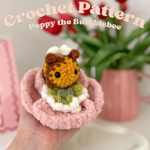 Könnte beinhalten: Eine Häkelanleitung mit dem Titel Crochet Pattern Poppy the Bumblebee, die ein kleines, flauschiges Hummelspielzeug aus braunem und gelbem Garn mit schwarzen Augen und grünem Körper zeigt, eingebettet in eine weiche, hellrosa, gerüschte Häkelblumenbasis.