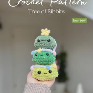 May include: A stack of three crocheted frog-like figures in shades of green and yellow, with black eyes and smiling faces. The top frog has a yellow crown. The image includes the text "Crochet Pattern Tree of Ribbits" and "low-sew".
