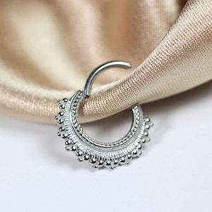 Puede incluir: Un anillo de septum plateado con un diseño de cuentas. El anillo con bisagras está parcialmente abierto, revelando el cierre. La joya se encuentra sobre un fondo de tela de satén beige y una superficie blanca.