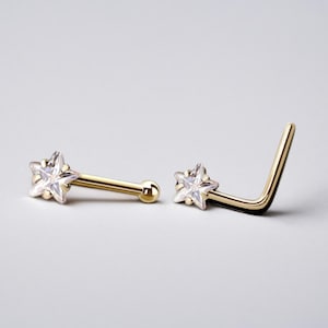 May include: Two gold nose rings with a star-shaped clear gemstone. One ring is straight and the other is L-shaped.