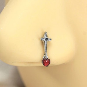 May include: A silver nose ring featuring a sword design with a small, dangling red heart-shaped charm. The jewelry is shown on a person's nose, with a close-up view highlighting the details of the piercing.