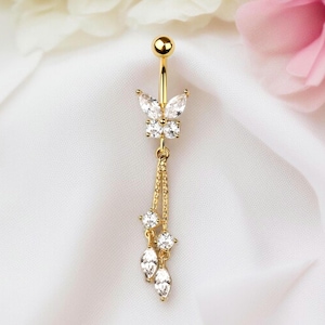 Puede incluir: Anillo para el ombligo dorado con un diseño de mariposa con cristales claros y brillantes. La mariposa está en la parte superior, con detalles de cristal colgantes. La joya está sobre un fondo de tela blanca, con flores rosas y blancas.