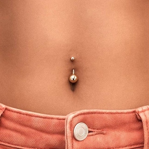 Puede incluir: Un piercing de ombligo dorado con una pequeña bola superior y una bola inferior más grande. El piercing está en el abdomen de una persona, con unos vaqueros color melocotón visibles en la parte inferior de la imagen.