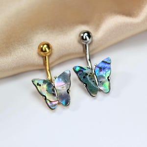 May include: Two belly button rings with butterfly pendants. One has a gold-colored barbell and the other has a silver-colored barbell. The butterfly pendants have iridescent, multi-colored wings. The jewelry is displayed on a white surface with a gold-colored fabric in the background.