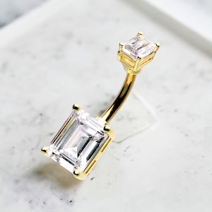 Op de afbeelding: Gouden navelring met twee heldere vierkante zirkonia-stenen.