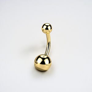 Puede incluir: Un piercing para el ombligo de color dorado con un diseño de barra curva. La joya presenta una pequeña bola en la parte superior y otra más grande en la inferior, ambas muy pulidas y reflectantes.