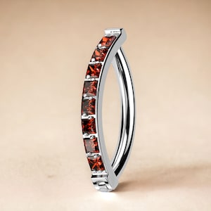 May include: A silver body jewelry ring with a curved shape and a row of red gemstones.