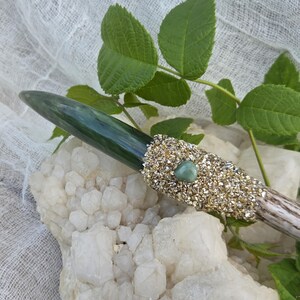 May include: A handcrafted wand with a green crystal tip and a decorative gold-colored band. The wand is adorned with a small green gemstone and rests on a white quartz crystal. Green leaves are in the background.