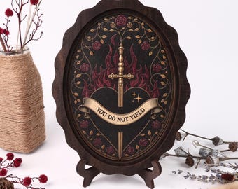 May include: An oval dark wood frame holds a black print featuring a central gold sword piercing a black heart, surrounded by red flames and roses. A gold banner across the heart displays the text YOU DO NOT YIELD in black capital letters.