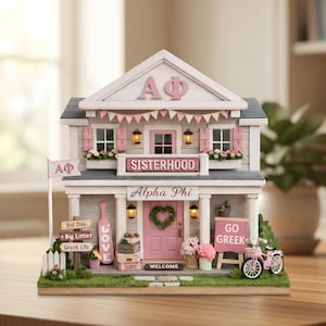 Puede incluir: Una casa en miniatura decorativa en rosa, blanco y gris, con los textos "Alpha Phi" y "Sisterhood". Incluye una pequeña bicicleta y letreros con "Go Greek" y "Welcome".