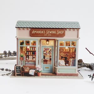 Puede incluir: Un modelo en miniatura de una tienda de costura titulada Amanda's Sewing Shop Est 1995. La estructura cuenta con un techo azul claro, una puerta central con una corona de flores y grandes escaparates que muestran estantes de telas e hilos.