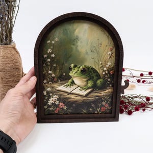 May include: A dark brown arched wooden frame containing a printed illustration of a green frog with large eyes sitting on the ground and reading an open book, surrounded by small white and red wildflowers in a soft, natural forest setting.