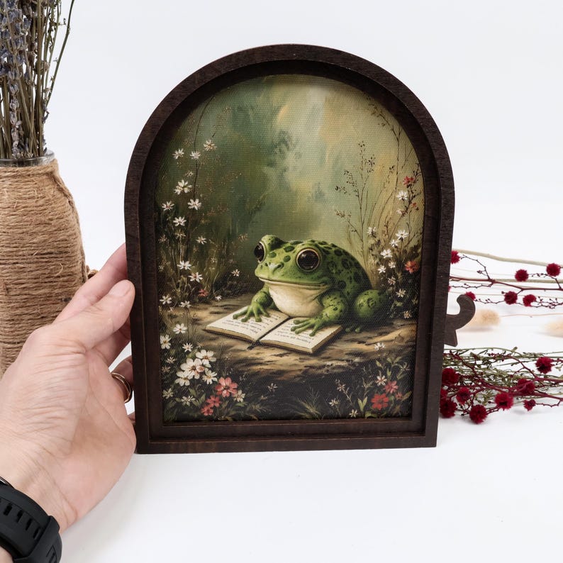 May include: A dark brown arched wooden frame containing a printed illustration of a green frog with large eyes sitting on the ground and reading an open book, surrounded by small white and red wildflowers in a soft, natural forest setting.