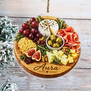 Könnte beinhalten: Ein rundes Holzornament mit einem aufgedruckten Motiv einer Wurst- und Käseplatte mit Trauben, Käse, Feigen, Oliven und Aufschnitt. Das Wort Aura ist unten in Schreibschrift eingraviert. Das Ornament hängt an einer dünnen Schnur.