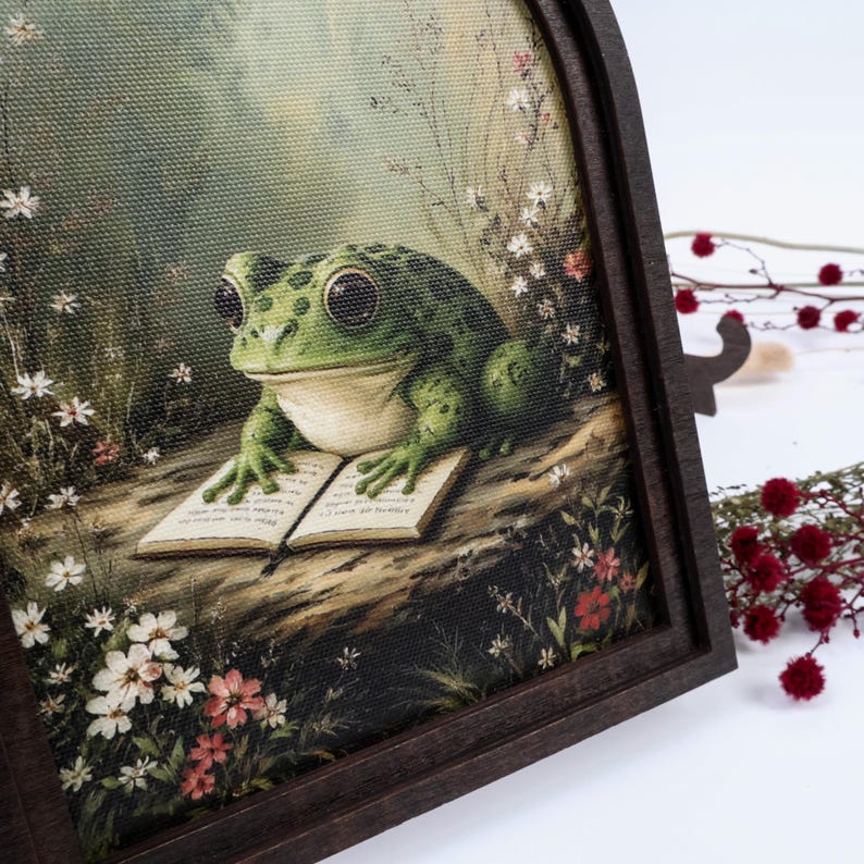 May include: A framed canvas print featuring a green frog with large eyes resting on a log while reading an open book. The scene is surrounded by small white and red wildflowers against a soft, muted background, all encased in a dark wood frame.