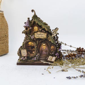 May include: A decorative fairy house sculpture featuring a tree-like structure with a purple roof, illuminated windows, and a sign reading Sophia's Fairy House. The base includes a sign with the text Dream Believe Imagine and an open book.