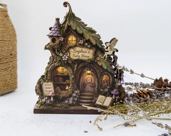 May include: A decorative fairy house sculpture featuring a tree-like structure with a purple roof, illuminated windows, and a sign reading Sophia's Fairy House. The base includes a sign with the text Dream Believe Imagine and an open book.
