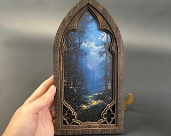 May include: A dark brown wooden frame with a pointed gothic arch shape containing a printed canvas image of a forest at night. The scene features tall trees, a crescent moon in a blue sky, and glowing yellow light patches on the forest floor.