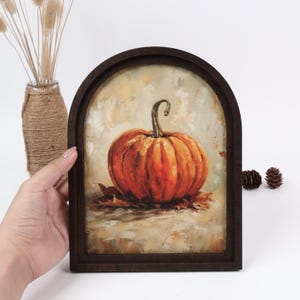 Pode incluir: Uma moldura de madeira arqueada contendo uma pintura a óleo de uma abóbora laranja com um caule curvo, repousando sobre folhas castanhas contra um fundo bege texturizado. A obra de arte apresenta pinceladas visíveis e tons quentes.