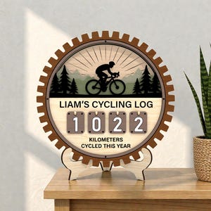 May include: Wooden cycling log in a gear-shaped frame. The centre shows a cyclist silhouette, trees, and the text "LIAM'S CYCLING LOG." It displays "1022" kilometres cycled this year. The log is supported by a wooden stand.