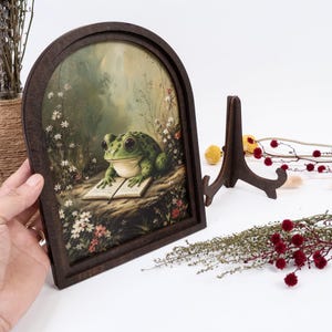May include: An arched wooden frame containing a printed illustration of a green frog with large eyes sitting on a rock and reading an open book. The scene is surrounded by small white and red flowers and tall grasses in a soft, muted forest setting.