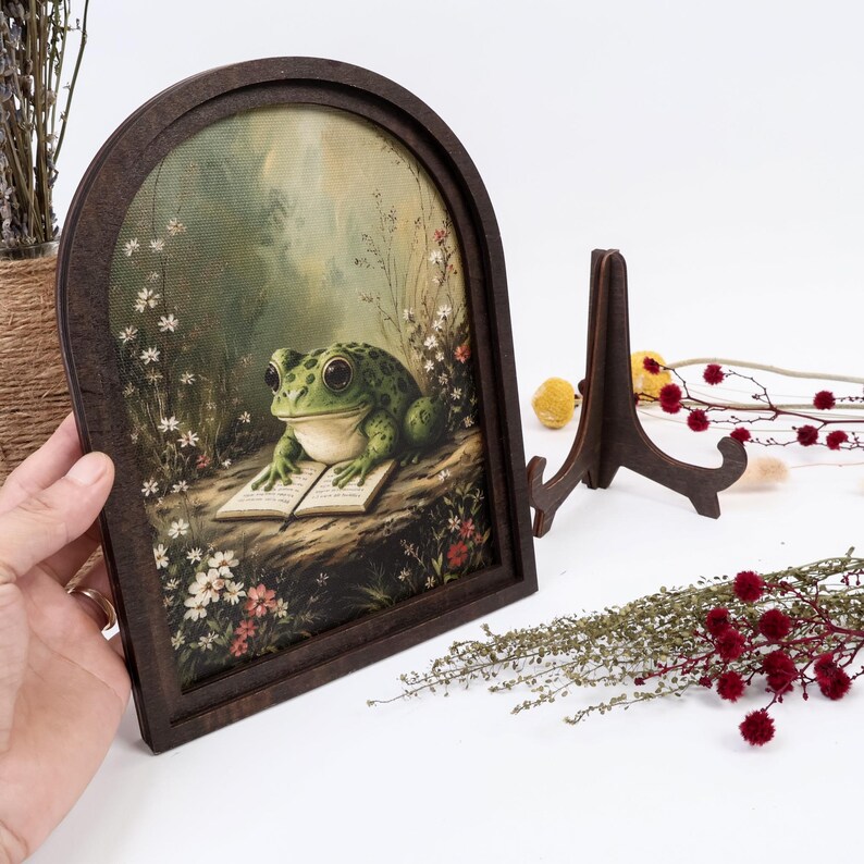 May include: An arched wooden frame containing a printed illustration of a green frog with large eyes sitting on a rock and reading an open book. The scene is surrounded by small white and red flowers and tall grasses in a soft, muted forest setting.