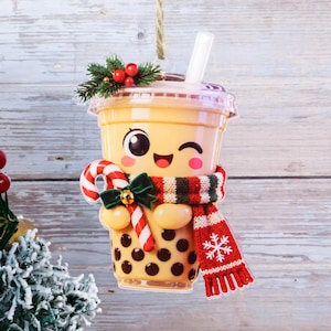 Könnte beinhalten: Ein Bubble-Tea-Becher-Anhänger mit beigefarbener Flüssigkeit, schwarzen Tapioka-Perlen und einem weißen Strohhalm. Der Becher zeigt ein Cartoon-Gesicht, eine rot-weiße Zuckerstange, eine grüne Schleife und einen rot-weiß-grünen Strickschal.