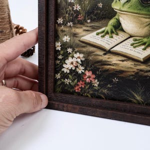 May include: A framed canvas print featuring a green frog with its front feet resting on an open book. The scene includes small white and pink wildflowers growing along the bottom left edge. The artwork is encased in a dark brown wooden frame.