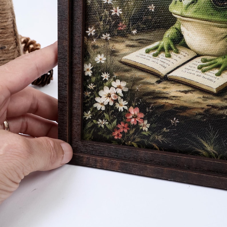 May include: A framed canvas print featuring a green frog with its front feet resting on an open book. The scene includes small white and pink wildflowers growing along the bottom left edge. The artwork is encased in a dark brown wooden frame.