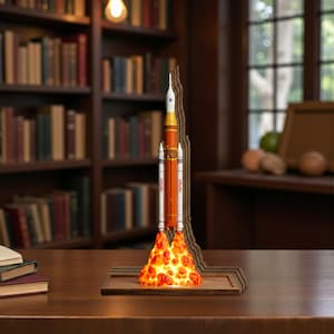 Könnte beinhalten: Dekorative Raketenschiff-Lampe mit orangefarbenen und weißen Akzenten, mit dem NASA-Logo. Die Lampe hat einen Holzsockel und einen feurigen, beleuchteten Effekt am Boden, der ein warmes Leuchten erzeugt. Die Rakete ist etwa 30 cm hoch.