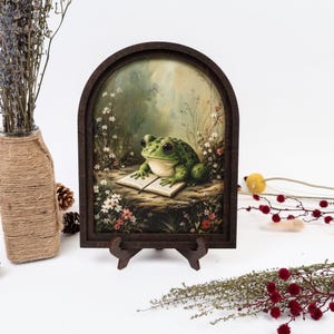 May include: An arched wooden frame containing a print of a green frog with large eyes sitting on a rock and reading an open book, surrounded by small white and red flowers in a meadow setting, supported by a small dark brown display stand.