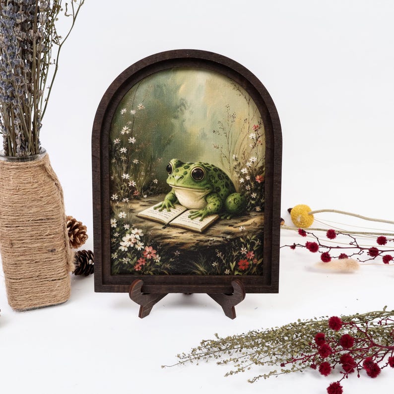 May include: An arched wooden frame containing a print of a green frog with large eyes sitting on a rock and reading an open book, surrounded by small white and red flowers in a meadow setting, supported by a small dark brown display stand.