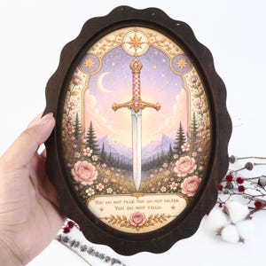 Könnte beinhalten: Ein ovaler Rahmen aus dunklem Holz mit einer Illustration eines Schwerts vor einer Berglandschaft mit rosa Blumen, einer Mondsichel und Sternen. Der Text lautet: You do not fear. You do not falter. You do not yield. Der Rahmen hat einen gewellten Rand.