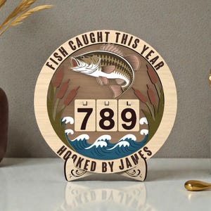 Puede incluir: Letrero de madera con temática de pesca con una ilustración de pez, el texto "FISH CAUGHT THIS YEAR" y el número "789". El letrero también incluye el texto "HOOKED BY JAMES" y olas decorativas.