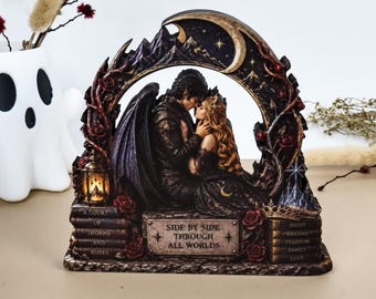 Puede incluir: Una escultura decorativa de resina que muestra una figura alada y una figura de pelo largo abrazadas bajo un arco de media luna. La base incluye pilas de libros con el texto A COURT OF THORNS AND ROSES y SIDE BY SIDE THROUGH ALL WORLDS.